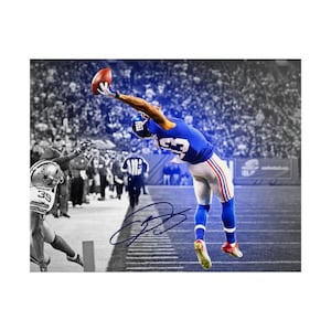 May include: A sports photograph featuring a football player in a blue jersey and white trousers leaping to catch a brown football. The player wears a blue helmet and blue socks. A signature is visible in blue ink across the centre of the image.