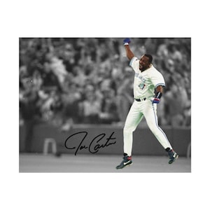 May include: A sports photograph featuring a baseball player in a white uniform with blue accents, jumping with one arm raised in celebration. The background is a blurred black and white crowd. The signature Joe Carter is printed in black ink.