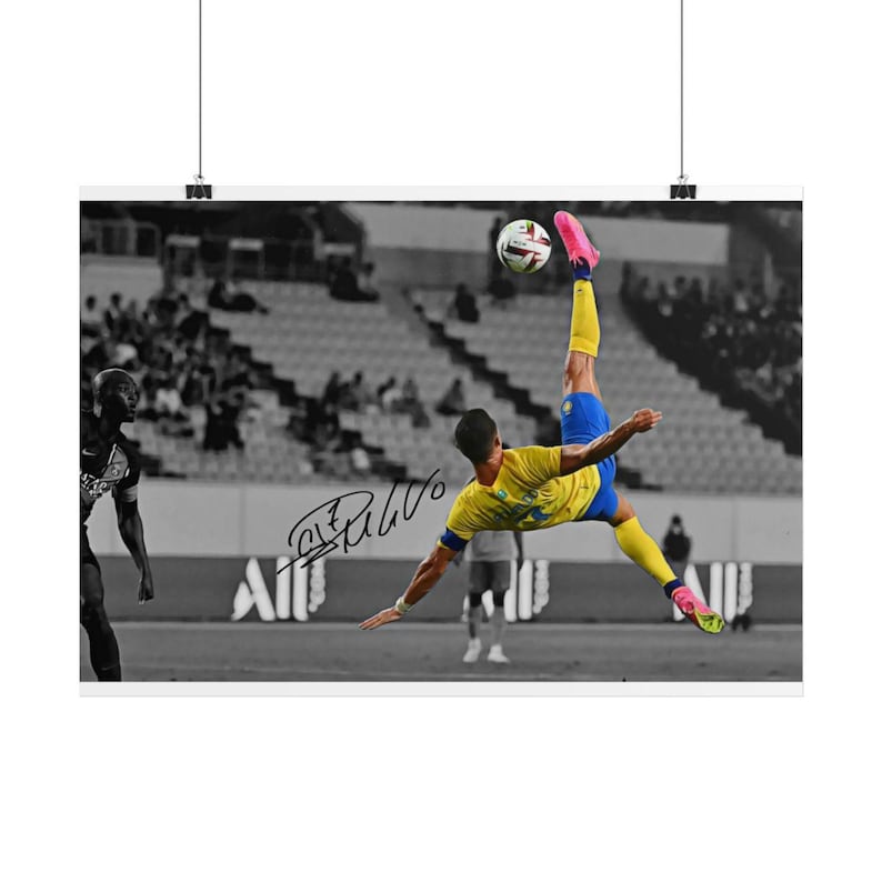 May include: A black and white sports poster featuring a soccer player in a yellow jersey and blue shorts performing a bicycle kick to strike a soccer ball. The player wears bright pink cleats and yellow socks. A black ink signature is visible.