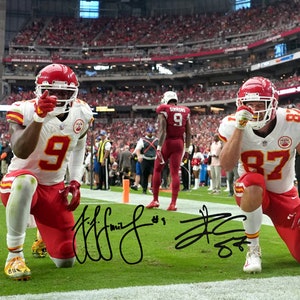 May include: Two football players in red and white uniforms kneel on the pitch, celebrating a touchdown. The player on the left is wearing number 9 and the player on the right is wearing number 87. Both players are holding their hands up in the air. The background shows a stadium full of fans.