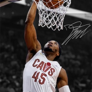 May include: A basketball player in a white jersey with "CAVS" and the number "45" in maroon, dunks a basketball into a net. The image is in color with a black and white background. A signature is in the upper right corner.