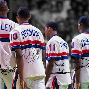 Op de afbeelding: Vier basketballspelers die witte shirts dragen met rode en blauwe strepen. De shirts hebben de namen en nummers van de spelers op de rug. De spelers staan ​​allemaal met hun gezicht naar voren en hebben hun armen langs hun zijden. De shirts zijn gesigneerd door de spelers.