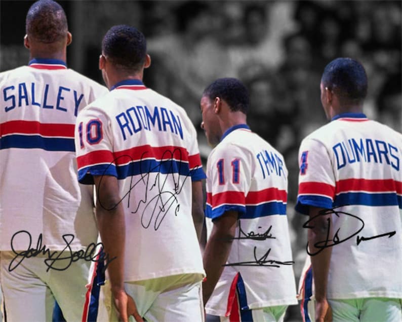 Op de afbeelding: Vier basketballspelers die witte shirts dragen met rode en blauwe strepen. De shirts hebben de namen en nummers van de spelers op de rug. De spelers staan ​​allemaal met hun gezicht naar voren en hebben hun armen langs hun zijden. De shirts zijn gesigneerd door de spelers.