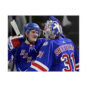 May include: A photograph of two hockey players in blue New York Rangers jerseys. One player wears a blue helmet and holds a hockey stick, while the other wears a goalie mask with red, white, and blue patterns. Both jerseys feature the name SHESTERKIN.