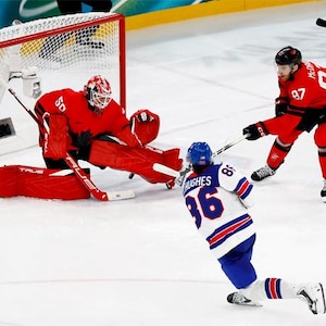 May include: A hockey game in progress, featuring players in red and white uniforms. A goalie in red gear defends the net, while two other players skate with sticks. The ice rink is white, and the net is red and white.