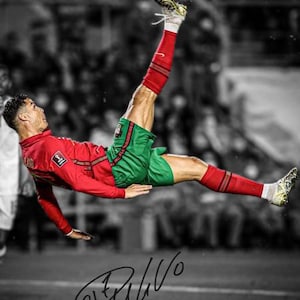 May include: A footballer in a red long-sleeved shirt and green shorts performs an overhead kick to strike a white, red and black football in mid-air. The player wears red socks and gold boots. A black signature is visible at the bottom of the image.