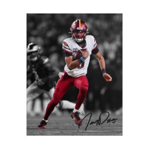 May include: A football player in a white jersey with burgundy accents and burgundy trousers, wearing a matching helmet, runs while holding a football. The background is blurred in black and white, and a black ink signature is visible in the bottom right.