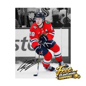 May include: A photograph of a hockey player wearing a red jersey with the number 98 and a black helmet, holding a hockey stick on the ice. The image features a black ink autograph signature across the bottom and a white background.