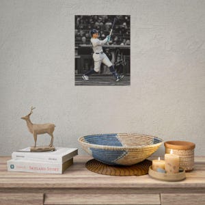 May include: A framed black and white photograph of a baseball player in a pinstriped uniform swinging a bat, featuring a signature on the image. Below, a wooden deer figurine sits on a book titled Skyland Story, next to a woven bowl and two candles.