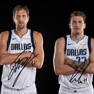 May include: Two basketball players wearing white jerseys with blue trim and the word "Dallas" on the front. The jerseys have the player's number on the front and their signatures on the front. The players are standing with their arms crossed.