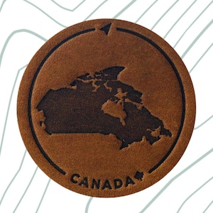 May include: A brown leather patch featuring an outline of Canada and the text "CANADA" in white. The patch has a black hook and loop backing.