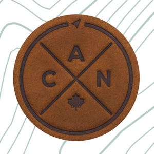 May include: A circular, brown leather patch featuring the letters "CAN" and a maple leaf symbol. The patch has a compass design and is shown with a black Velcro hook backing. The text "Velcro Hook Backing" is also visible.