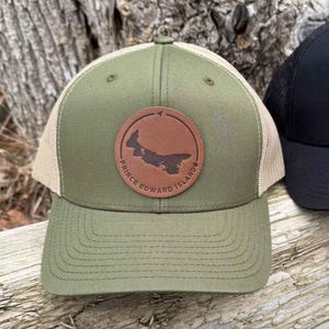Peut inclure: Deux casquettes de camionneur avec des patchs en cuir. La casquette verte a un patch brun avec le contour de l'Île-du-Prince-Édouard et le texte "Île-du-Prince-Édouard". La casquette noire a un patch brun avec le contour du Manitoba et le texte "Manitoba".