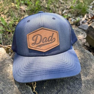May include: A navy blue trucker hat with a curved brim. The hat features a brown leather patch with the word "Dad" in a script font. The hat has a mesh back and a snap closure.