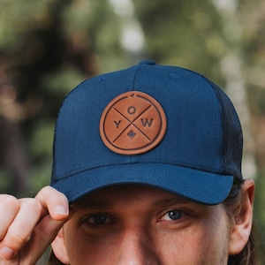 May include: A navy blue trucker hat with a brown leather patch featuring the letters "YOW" and a Canadian maple leaf. The hat has a black mesh back.
