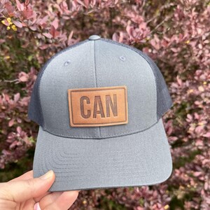 May include: Gray trucker hat with a dark gray mesh back and a curved brim. A rectangular brown leather patch on the front displays the word "CAN" in capital letters. The hat is set against a backdrop of reddish-purple foliage.