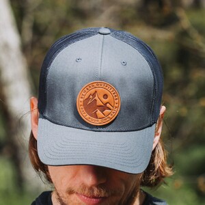 May include: A navy blue and black mesh trucker hat with a brown leather patch that says "Byward Outfitters Craft Your Own Path".
