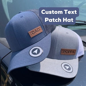 May include: Two trucker hats with custom leather patches. The blue hat has a brown patch that says "DAD" and the grey hat has a brown patch that says "POPPA".