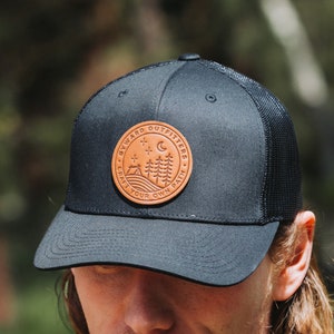 Peut inclure: Casquette de camionneur noire et marron avec un patch en cuir qui dit "Byward Outfitters - Craft Your Own Path" avec une lune croissante et des pins.