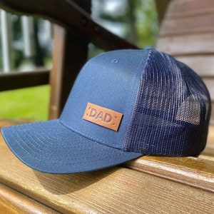 Peut inclure: Un chapeau de camionneur bleu marine avec un patch en cuir marron qui dit "DAD" sur le devant.