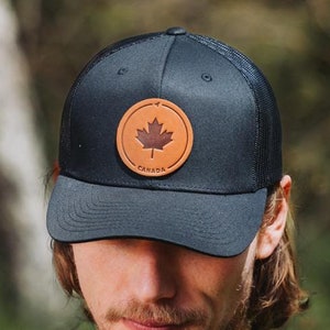 May include: A black mesh trucker hat with a brown leather patch featuring a maple leaf and the word "Canada".