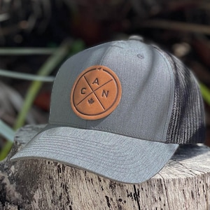 Peut inclure: Casquette de baseball grise avec un dos en filet noir. Le chapeau comporte un écusson en cuir marron avec les lettres "CAN" et un emblème de feuille d'érable. Le chapeau est posé sur une surface en bois.