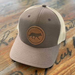 May include: A brown and beige trucker hat with a brown leather patch featuring a silhouette of a wolf. The patch is circular and has a white arrow pointing to the right.