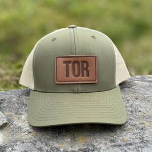 May include: An olive green trucker cap with a tan mesh back and a brown leather patch. The patch displays the word "TOR" in capital letters. The hat is resting on a rock with a blurred green background.