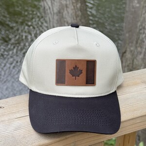 May include: A cream-colored baseball cap with a black brim and a rectangular brown leather patch on the front. The patch features a dark brown Canadian maple leaf emblem centered within a design that mimics the Canadian flag's vertical stripes.