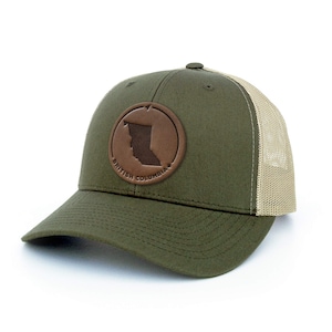 May include: A green and tan trucker hat with a brown leather patch featuring the outline of British Columbia. The patch has the text "BRITISH COLUMBIA" around the outline.