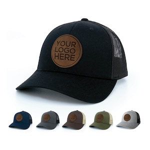 Peut inclure: Un chapeau de camionneur noir avec un patch en cuir marron qui dit "YOUR LOGO HERE". Le chapeau a une face avant noire et un dos en maille noire. Il y a aussi cinq autres chapeaux de différentes couleurs : bleu marine, gris, marron, vert et gris.