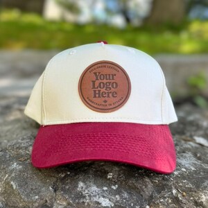May include: A cream and burgundy baseball cap with a leather patch. The patch features the text "Your Logo Here" and "Handcrafted in Ottawa". The cap is designed for outdoor use and is made from a robust material. The brim is burgundy.