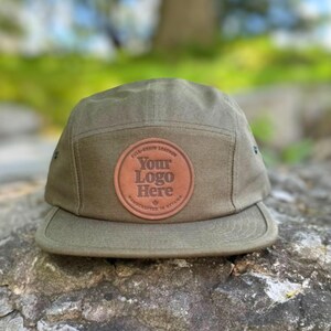 May include: An olive green five-panel cap with a brown leather patch. The patch displays the text "Your Logo Here" in a circular design. The cap is made of a durable fabric and has a flat peak. The text "Handcrafted in Ottawa" is also visible.