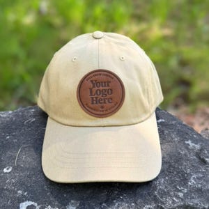 May include: A beige baseball cap with a curved peak. A brown leather patch on the front features the text "Your Logo Here" in white, with surrounding text. The cap is resting on a rock.
