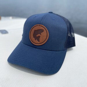May include: A navy blue trucker hat with a brown leather patch featuring a silhouette of a fish. The hat has a mesh back.