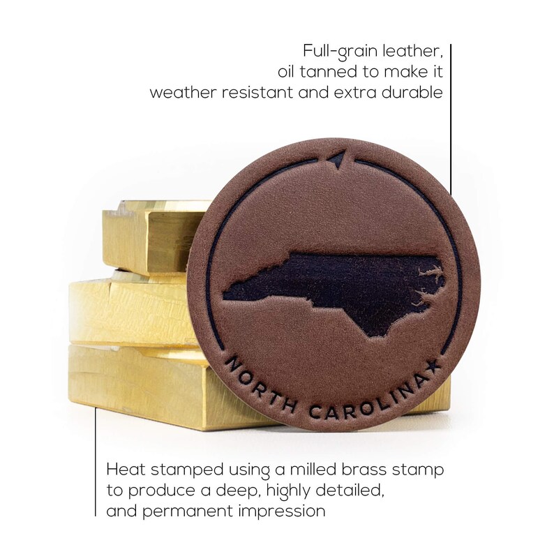 May include: A brown leather patch with a black outline and the state of North Carolina in black inside. The patch is heat-stamped with the text "NORTH CAROLINA*" and the words "Full-grain leather, oil tanned to make it weather resistant and extra durable" are written above the patch. The patch is sitting on top of a stack of brass blocks.