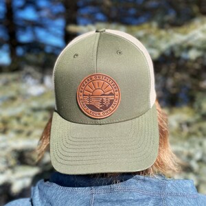 Peut inclure: Un chapeau de camionneur vert et blanc avec un patch en cuir marron qui dit "Byward Outfitters Craft Your Own Path" avec un design de soleil et d'arbres.
