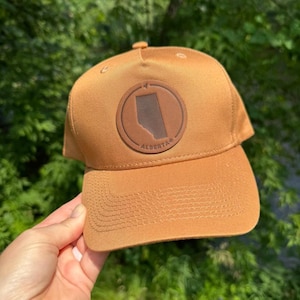 May include: A tan baseball cap featuring a circular brown patch on the front panel. The patch displays a silhouette of the province of Alberta and the text ALBERTA. The cap has a structured crown, a curved brim with stitching, and a button on top.