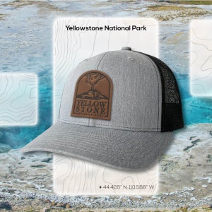 May include: A gray trucker hat with a black mesh back and a curved brim. The hat features a brown leather patch with the words "Yellowstone National Park" and a tree graphic. The text "Yellowstone National Park" is also visible.