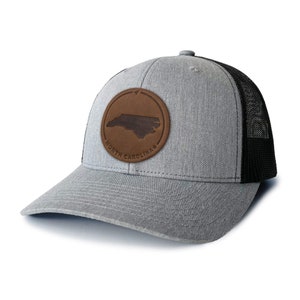 May include: A grey and black trucker hat with a brown leather patch featuring the outline of North Carolina and the text "NORTH CAROLINA".