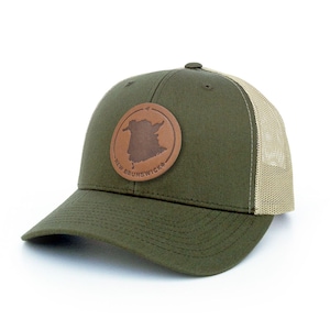 May include: Olive green baseball cap with a tan mesh back. A circular brown leather patch on the front displays the outline of New Brunswick, with the text "NEW BRUNSWICK" around the edge. The cap has a curved brim and a button on top.