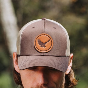 May include: A brown and beige trucker hat with a brown leather patch featuring a silhouette of a flying eagle. The patch is centered on the front of the hat.
