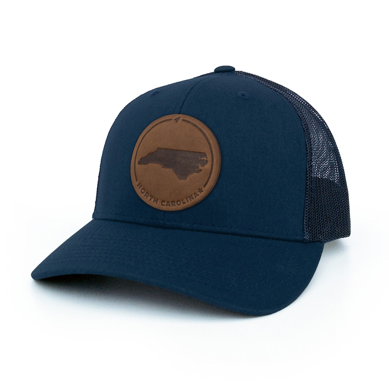 May include: A navy blue trucker hat with a brown leather patch featuring the outline of North Carolina and the text "NORTH CAROLINA".