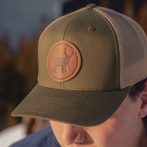 May include: A green and brown trucker hat with a leather patch featuring a silhouette of a reindeer. The hat has a mesh back.