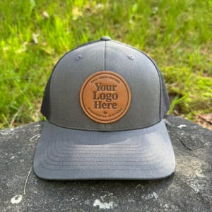 Peut inclure: Casquette de camionneur grise avec un dos en filet noir et une visière incurvée. Un écusson rond en cuir marron sur le devant affiche le texte "Your Logo Here" et "Full-Grain Leather, Handcrafted in Ottawa".