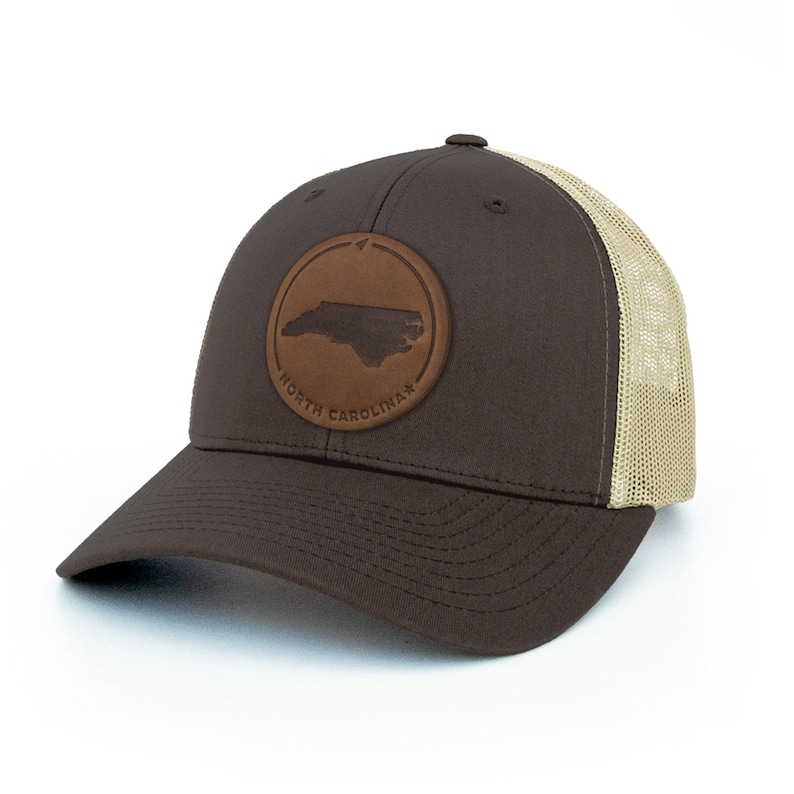 May include: A brown and tan trucker hat with a brown leather patch featuring the outline of North Carolina. The patch has the words "North Carolina" written around the state.