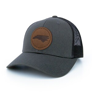 May include: A charcoal grey trucker hat with a black mesh back. The hat features a brown leather patch with the outline of North Carolina and the text "North Carolina".