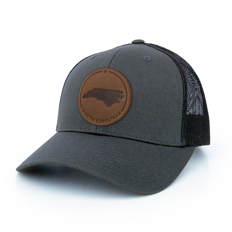 May include: A charcoal grey trucker hat with a black mesh back. The hat features a brown leather patch with the outline of North Carolina and the text "North Carolina".