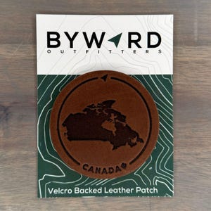 May include: A brown leather patch featuring an outline of Canada and the text "CANADA" in a white font. The patch is attached to a white card with the text "BYWARD OUTFITTERS" and "Velcro Backed Leather Patch".