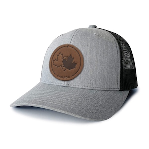 May include: A gray and black trucker hat with a brown leather patch featuring a maple leaf and the word "CANADA".